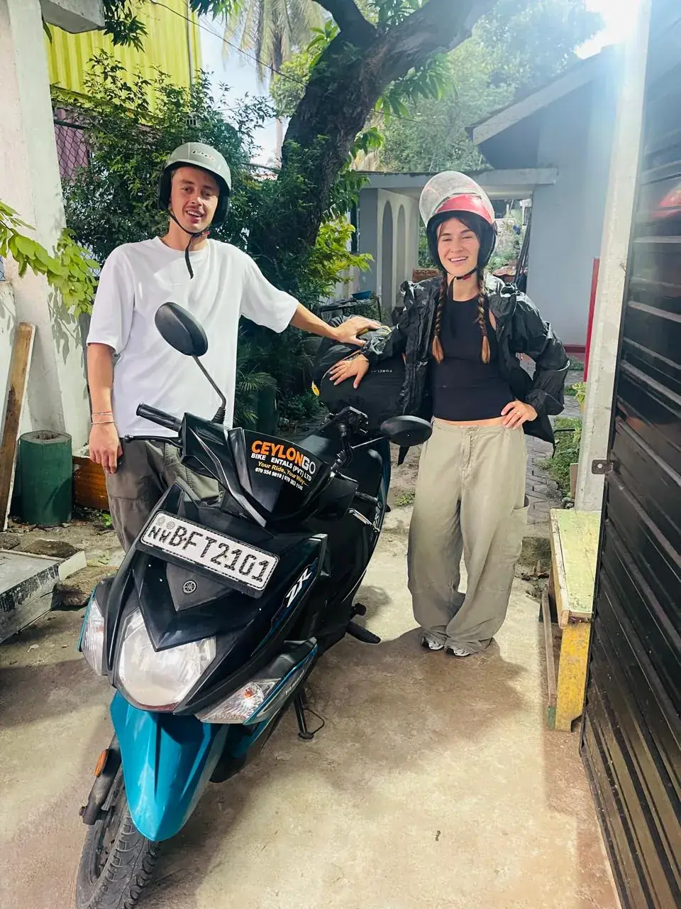 Couple smiling with helmets beside a CeylonGo branded Yamaha Ray ZR